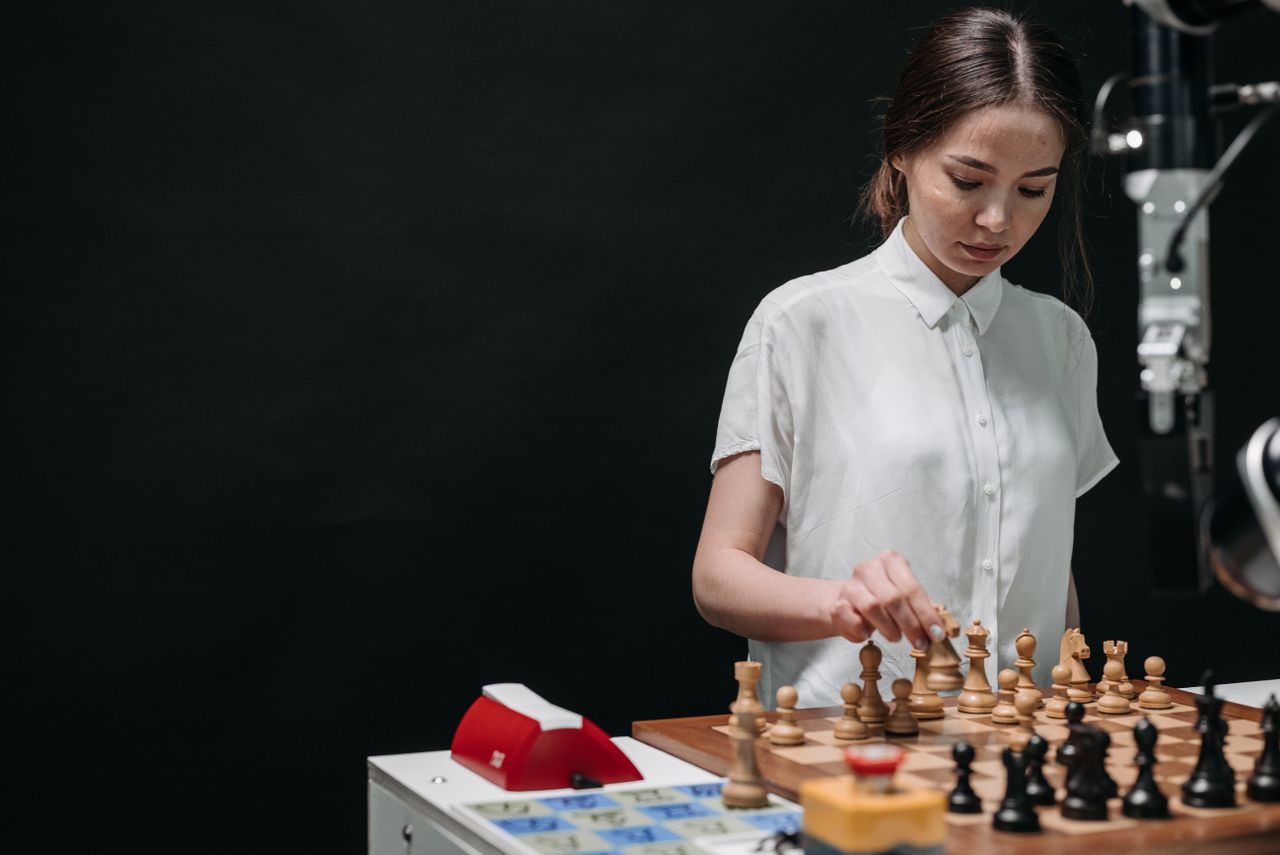 A woman plays chess against an articulated robot, symbolizing AI and human collaboration.